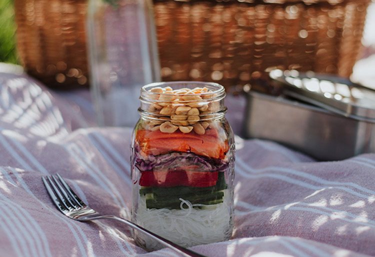 Ein Glasnudel-Salat im Glas steht auf einer Picknickdecke.