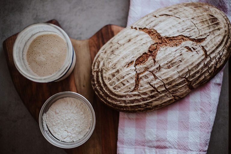 Selbst gebackenes Sauerteigbrot liegt neben Mehl und Sauerteig-Starter auf einem Schneidebrett.