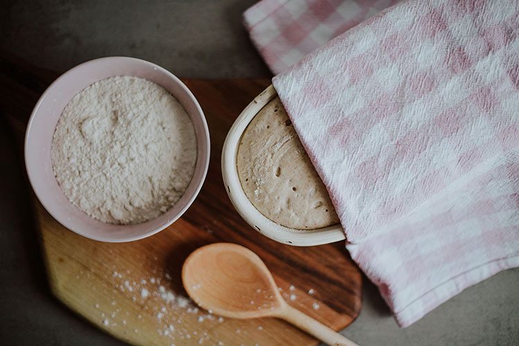 Ein Sauerteigbrot geht in einem Gärkörbchen mit einem Geschirrtuch abgedeckt auf.